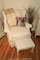 Wingback chair with matching foot stool, bunny pillow, and wool throw arranged together in corner of room with wood floor and neutral walls.