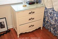 Full view of the small cream dresser with glass top showing three drawers and ornate handles.