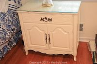 Front view of white nightstand showing glass top, drawer with bronze handle, and two door cabinet with matching handles.