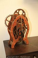 Front view of hand carved wooden clock showing wooden gears and pendulum on a wooden table.
