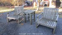 Photo showing two teak armchairs and a teak side table positioned outdoors on gravel ground with natural background.