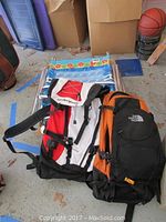 Two backpacks (Dakine and The North Face) resting on two folding beach chairs with floral wave patterned fabric, items arranged on the floor.