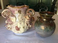 Photo showing two ceramic vases placed on a table: one ornate with floral design and sculptural handles, the other a simpler green ceramic vase.
