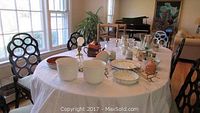 Wide view of dining table with ceramic items, glassware, and lamp set up to show lot contents.