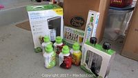 Photo showing boxed Soda Stream Genesis G100 soda maker, several 500ml syrup bottles with different flavors including Lemon Lime, Root Beer, Lemon Ice Tea, and Cola, and a large Soda Club shipping box.