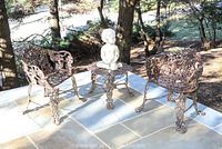 Outdoor wrought iron table and two matching wrought iron chairs placed on stone patio outdoors with natural background of trees and grass