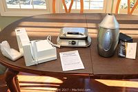 All three kitchen appliances displayed on a wooden table with sunlight: white Toastmaster deli slicer, Sharper Image silver wine cooler, and Cuisinart waffle maker with control dial.