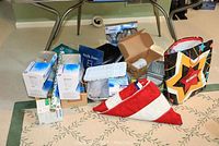 Overview of the lot showing the folded large American flag, boxes of light bulbs, greeting cards, shelf liner, metal containers, paper plate holders, gift bags and assorted household items arranged on the floor and table.