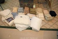 Overview of linens, sheets, and bedding items including heated blanket and mattress protectors, spread and folded on carpeted floor.
