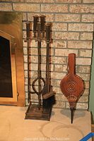 Full view of the metal fireplace tool set with stand next to a brick fireplace and the leather bellows upright on the floor.