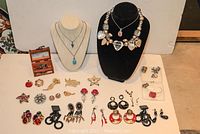Full lot displaying assorted earrings, brooches, small jewel box, and necklaces arranged on and around necklace busts and flat on table