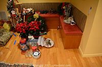 Wide shot showing corner seating area with multiple Christmas wreaths, red artificial poinsettias in pots, cookie tins, and various holiday items arranged on floor and seating bench.