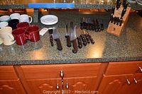 Countertop display showing Henckels knife block with knives (missing shears), Farberware steak knives, assorted coffee mugs, and some white plates.