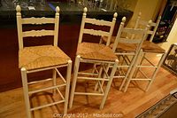 Four wooden bar stools with rush seats placed in front of a bar counter, showing design and finish wear.