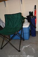 Photo showing green folding chair, blue Coleman cooler with white lid, green insulated bag, and multiple golf umbrellas along with a blue nylon folding chair in carry bag.