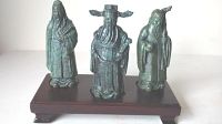 Front view of three metal statues depicting Asian gods of wealth, long life, and wisdom on polished wooden base showing detailed features and patina.