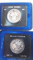 Close-up of both Canadian silver dollar coins in respective blue clam shell display cases, showing British Columbia and Manitoba commemorative designs and dates.