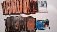 Photo shows two piles of trading cards: one pile of Magic: The Gathering cards including a face up card and an event deck card, and another pile of Yu-Gi-Oh! cards with card backs showing.