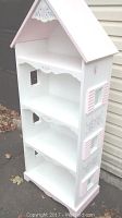 Front and angled view of a pink and white dollhouse-shaped bookcase with three shelves and a peaked roof.