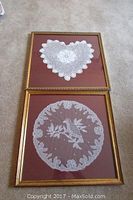 Two framed lace artworks on carpet: top is crocheted heart, bottom is circular bird bobbin lace.