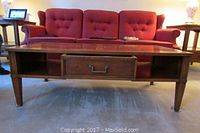 Full view of wooden coffee table with central drawer, tapered legs, and open shelves, placed in front of a red couch.