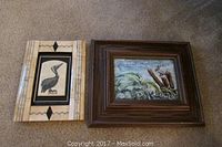 Two framed art pieces on carpet: on left, pelican hand engraved tile in cow bone frame; on right, watercolor nature scene framed in wood.