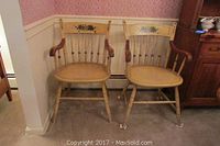 Pair of vintage wooden arm chairs with painted floral designs on the backrests placed side by side on carpeted floor against wallpapered wall.