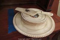 Stacked dessert dishes showing six small plates and one large cake plate with 22kt gold rimmed scalloped edges and decorative center images. Cake plate has a chip.