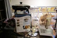 Photo showing the Rival Crock-Pot slow cooker in original box, Classic Chef food processor, Oster blender, and black electric skillet with glass lid on a wooden table.