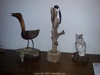 Photo showing all three carved wooden birds on a shelf: the Downy Woodpecker on tree branch in the center, a curved-neck bird on the left, and an owl on the right.