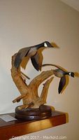 Side view of the Frank Muller carved geese sculpture showing the geese attached to a natural wood branch base on a wooden round platform.