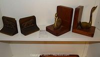 Both sets of bookends displayed on a shelf, one set with brass lions attached to dark metal bases and the other set with brass swans on wooden blocks.