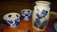 Three hand thrown pottery pieces by Eldreth on wooden surface: one tall vase with blue floral design and two votive holders with heart cutouts and blue decorative paint.