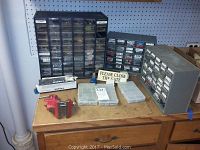 Three racks with labeled compartments filled with assorted screws, nails, picture hangers, o-rings, clamps, and other small hardware on a wood surface