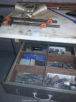 Top view of wooden drawer organizer with a pair of orange-handled bar clamps on top, and assorted drill bits, Allen wrenches, and other tools inside compartments.