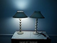 Pair of vintage cream metal tole painted lamps on top of a dresser with white linen scarf underneath. Both lamps are turned on showing their warm glow and some wear on bases and shades.
