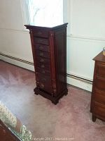 Full view of tall wooden jewelry box with decorative carved sides and brass hardware, standing on carpeted floor.