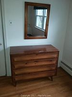 Medium shot of mid-century wooden dresser and wall mirror in a room with white walls and wood flooring.