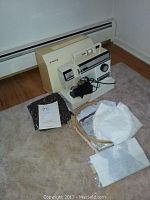Front view of sewing machine with power cord, fabric pieces, wooden embroidery hoop, and additional supplies on carpeted floor.