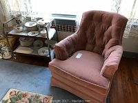 Rocking armchair in dusty rose color placed indoors near window and shelf, showing fabric upholstery, tufted backrest, and padded arms.