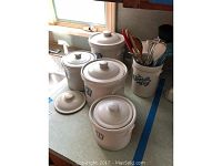 Six white stoneware canisters with lids, arranged on a kitchen counter near window light. Five of the canisters display blue numbers in vintage decal style: 1, 2, 3, 5, and 7, the sixth one does not display a number but appears similar in design.