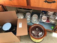 Brown Crockpot slow cooker with glass lid, stacked ceramic bowls and cups with floral designs, boxes possibly containing cookware.