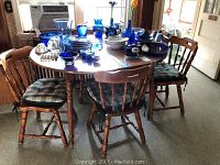 Vintage wooden kitchen table with one leaf and five wooden chairs with blue and white cushioned seats occupying a room with carpet flooring under natural light.