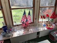 Eight glass vases, three U.S. flags, and other decorative items arranged on a window sill showing variety of clear glass vases and flags.