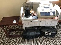 White bench with two drawers shown holding various household electronics, next to small brown snack table on striped carpet.
