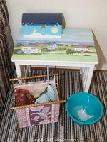 Photo showing the small painted side table with a beach scene on top, small wooden box decorated with sun, moon, stars on top of the table, fabric yarn holder basket with yarn and knitting needles next to table, teal bowl on the floor beside basket.