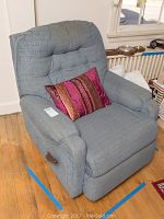 Blue fabric recliner chair shown in a room, featuring a tufted backrest and a colorful pink-red decorative pillow. Chair has a reclining lever on the right side and appears aged and used.
