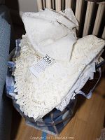Bag filled with assorted white and cream lace and fabric table linens including doilies, placemats, and napkins piled loosely.