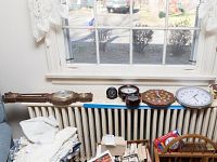 View of multiple clocks and barometers arranged on a window sill and radiator in front of a window. Includes wooden barometer clock with humidity scale and thermometer, wooden octagonal clock, white-faced quartz wall clock, two small black round clocks/gauges, small metal gauge, and wooden horizontal thermometer/barometer.
