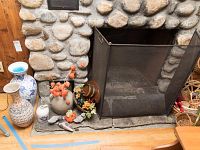 Photo shows stone fireplace with a black mesh fireplace screen; on hearth are multiple pottery and ceramic pieces including a large blue and white vase, white and blue lamp base, and neutral color vases. Several sculpted duck figurines and a rustic wooden grape and wine barrel fixture are displayed.